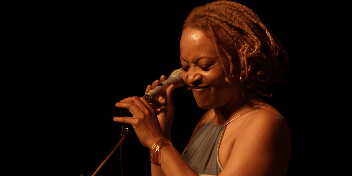A woman with dreadlocks smiles while holding a microphone and singing on stage