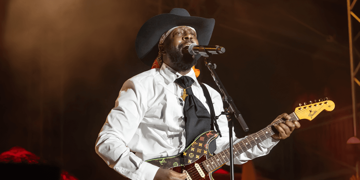 A musician wearing a cowboy hat performs on stage with an electric guitar