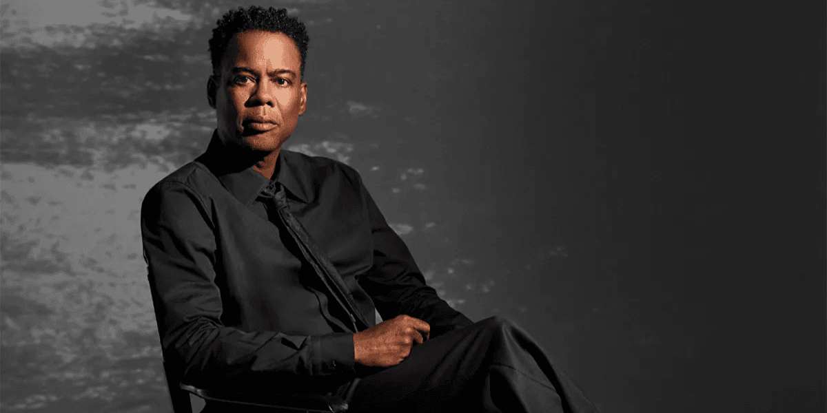 A man in a black shirt and tie seated against a muted background
