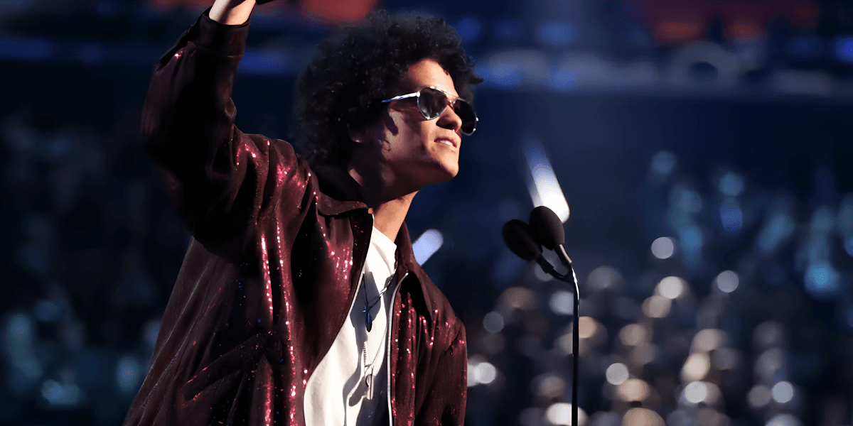 A man with curly hair wearing sunglasses gestures while speaking on stage