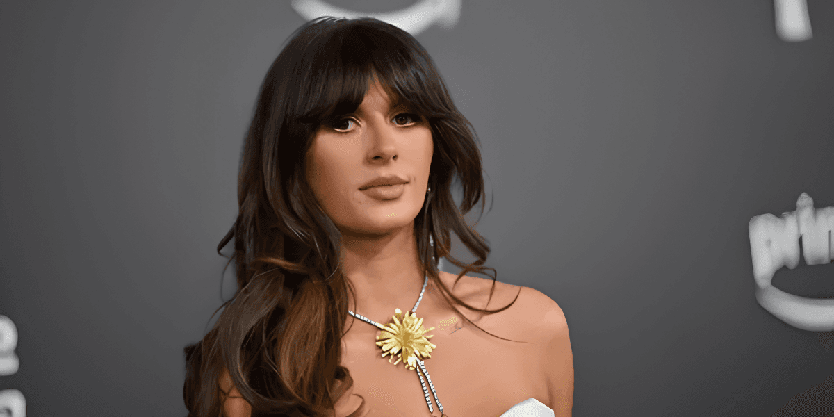 A woman with long hair wearing a white dress and floral necklace, posing seriously