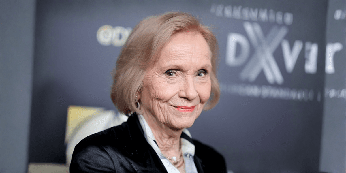 Elderly woman with short blonde hair smiling at an event backdrop