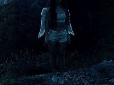 Woman with angel wings standing barefoot outdoors at dusk in a dark, cinematic forest landscape.