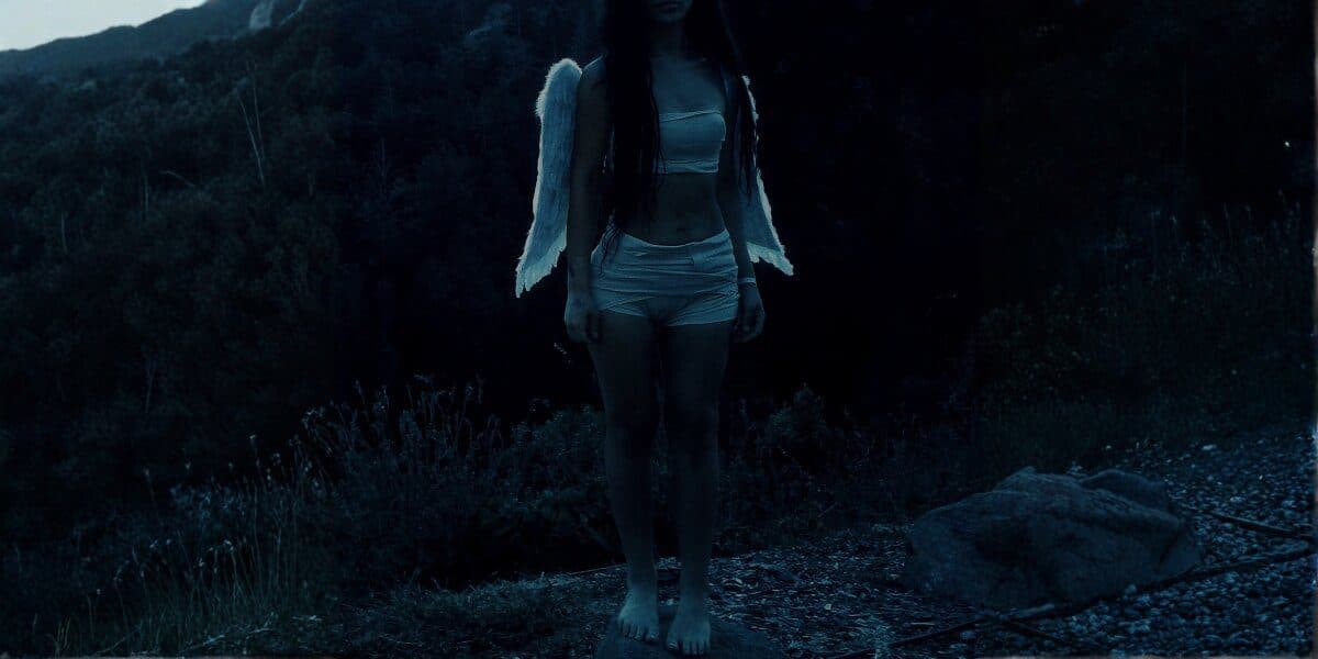 Woman with angel wings standing barefoot outdoors at dusk in a dark, cinematic forest landscape.