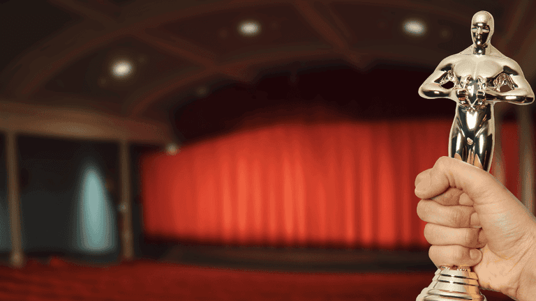 Hand holding a gold award statuette in front of a theater stage with red curtains.