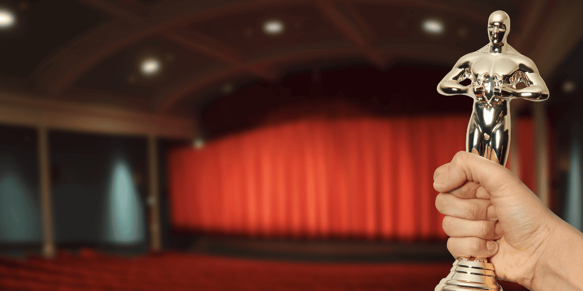 Hand holding a gold award statuette in front of a theater stage with red curtains.