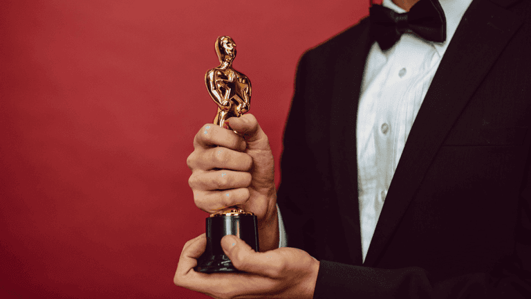 Person in a tuxedo holding a gold award statuette against a red background.