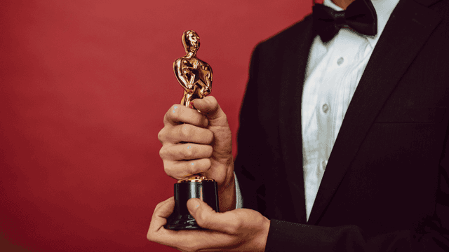 Person in a tuxedo holding a gold award statuette against a red background.