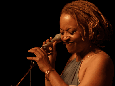 A woman with dreadlocks smiles while holding a microphone and singing on stage