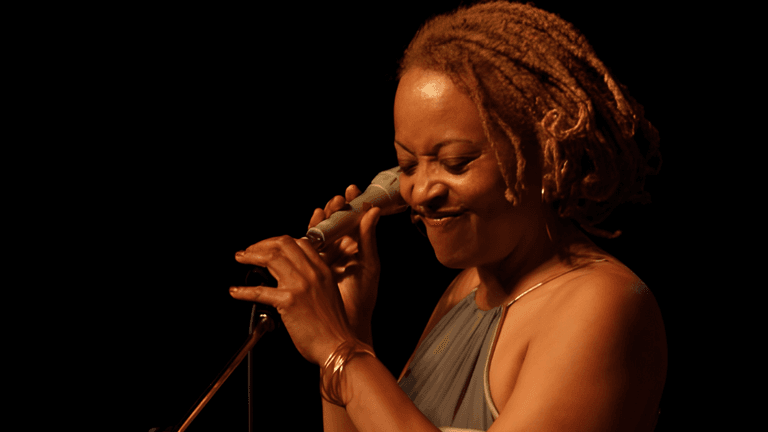 A woman with dreadlocks smiles while holding a microphone and singing on stage