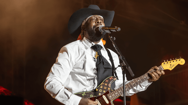 A musician wearing a cowboy hat performs on stage with an electric guitar