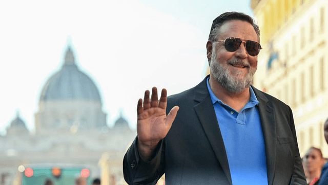 A smiling man in sunglasses waves, with St. Peter's Basilica in the background