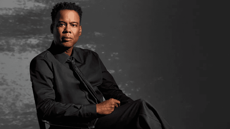 A man in a black shirt and tie seated against a muted background