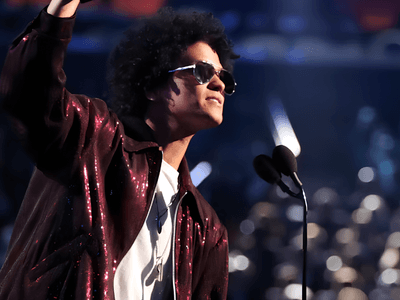 A man with curly hair wearing sunglasses gestures while speaking on stage