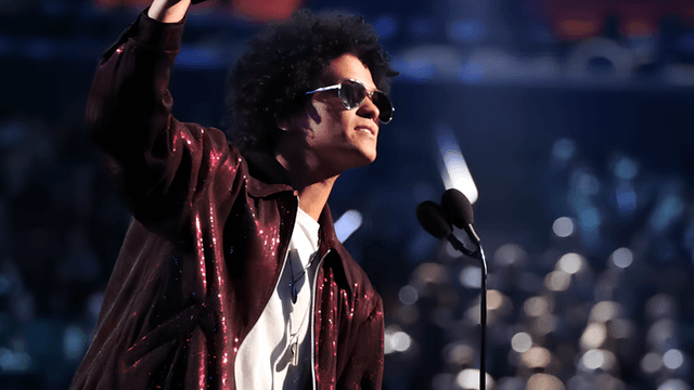 A man with curly hair wearing sunglasses gestures while speaking on stage