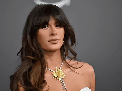 A woman with long hair wearing a white dress and floral necklace, posing seriously