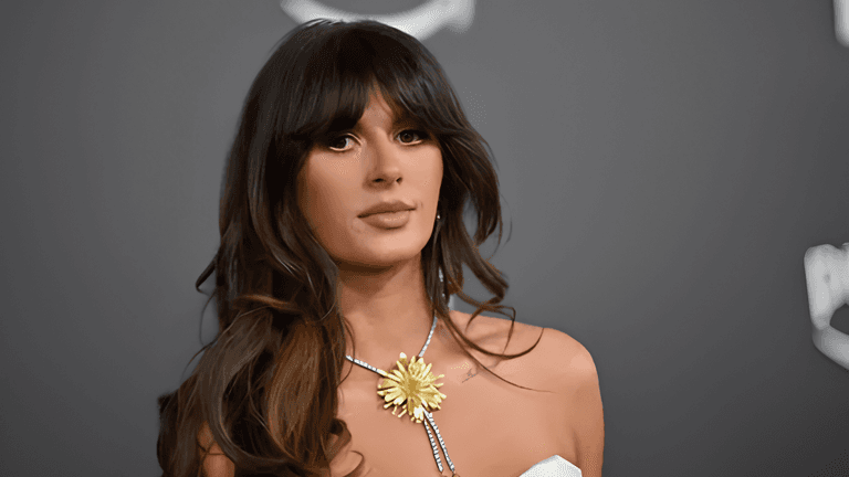 A woman with long hair wearing a white dress and floral necklace, posing seriously