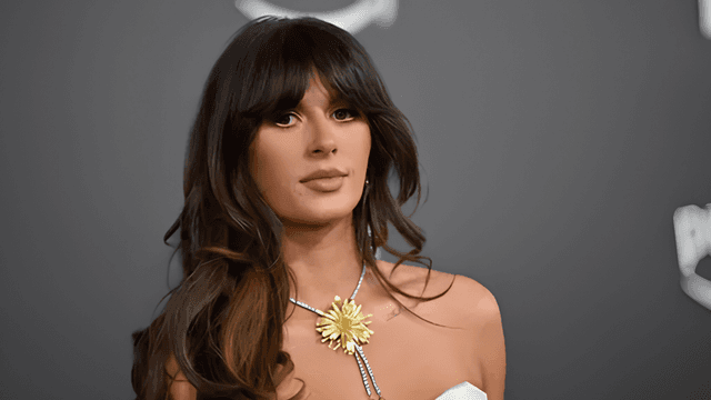 A woman with long hair wearing a white dress and floral necklace, posing seriously