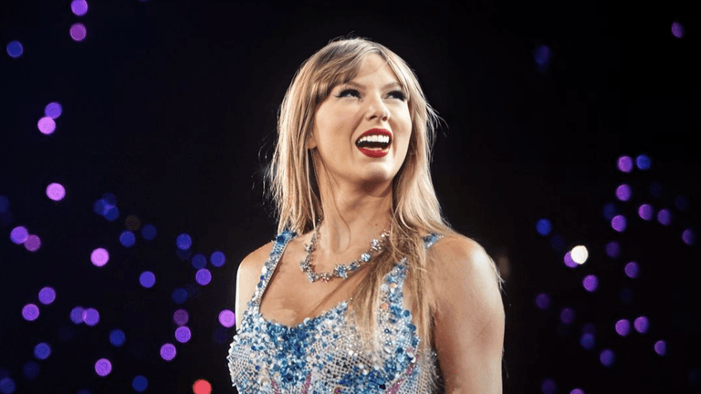 A performer in a sparkling outfit smiles brightly on stage with vibrant lights behind.