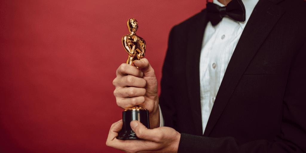 Emmy Buzz Continues as Television’s Biggest Nominees Build Momentum Ahead of Awards Season Person in a tuxedo holding a gold award statuette against a red background.